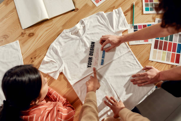 Top view of creative people trying on stickers with text, while discussing logo and design of T-shirt. Young man and women working together at custom T-shirt, clothing printing company. Cropped shot